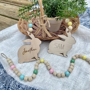 K&ouml;nnte beinhalten: Zwei Holzhasenformen mit den Namen "Jonathan" und "SAM" darauf geschrieben. Die Hasen h&auml;ngen an einer Girlande aus Holzperlen mit pastellblauen, rosa und gr&uuml;nen Akzenten.