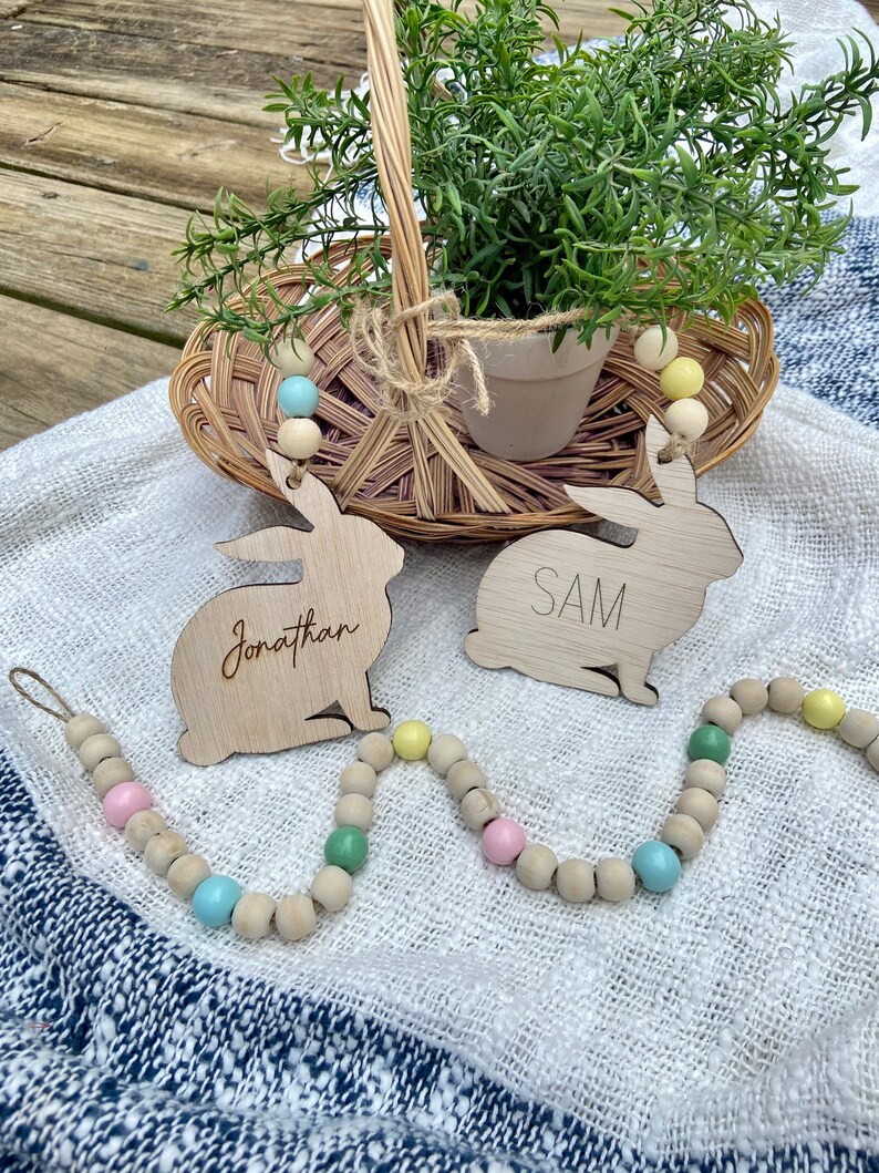 K&ouml;nnte beinhalten: Zwei Holzhasenformen mit den Namen "Jonathan" und "SAM" darauf geschrieben. Die Hasen h&auml;ngen an einer Girlande aus Holzperlen mit pastellblauen, rosa und gr&uuml;nen Akzenten.