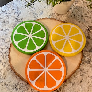 Puede incluir: Tres posavasos de madera con forma de rodajas de c&iacute;tricos. Los posavasos est&aacute;n pintados de verde, amarillo y naranja, con contornos blancos. Los posavasos est&aacute;n dispuestos sobre una rodaja de madera.