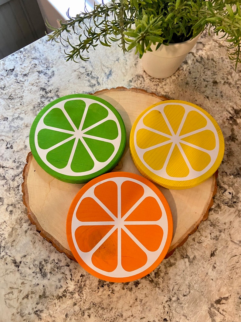Puede incluir: Tres posavasos de madera con forma de rodajas de c&iacute;tricos. Los posavasos est&aacute;n pintados de verde, amarillo y naranja, con contornos blancos. Los posavasos est&aacute;n dispuestos sobre una rodaja de madera.