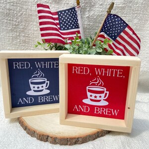 Pu&ograve; includere: Due cartelli in legno con il testo "Red, White, and Brew" e un'immagine di una tazza di caff&egrave;. I cartelli sono dipinti in rosso, bianco e blu. I cartelli sono appoggiati su una fetta di legno.