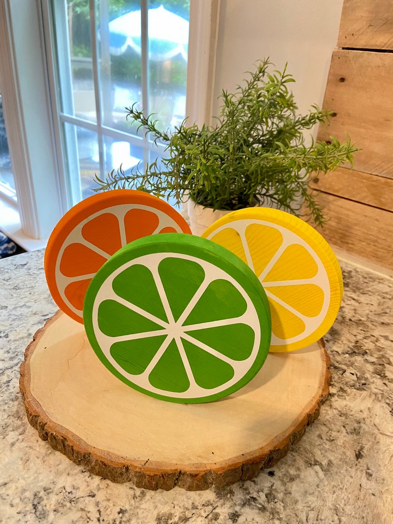 Puede incluir: Tres rodajas de fruta de madera pintadas en naranja, verde y amarillo, que se asemejan a una naranja, una lima y un lim&oacute;n. Las rodajas est&aacute;n dispuestas sobre una rodaja de madera de un tronco de &aacute;rbol.