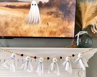 May include: A decorative garland featuring six small white fabric ghosts with black eyes, tied with black and white twine, strung along a strand of alternating natural wood and black beads, designed for hanging display on a fireplace mantel.