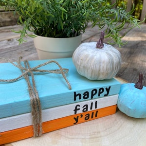 Puede incluir: Tres bloques de madera apilados pintados en azul, blanco y naranja con las palabras "happy fall y'all" escritas en negro. Los bloques est&aacute;n atados con cuerda y est&aacute;n sobre una rodaja de madera con dos peque&ntilde;as calabazas blancas y azules.