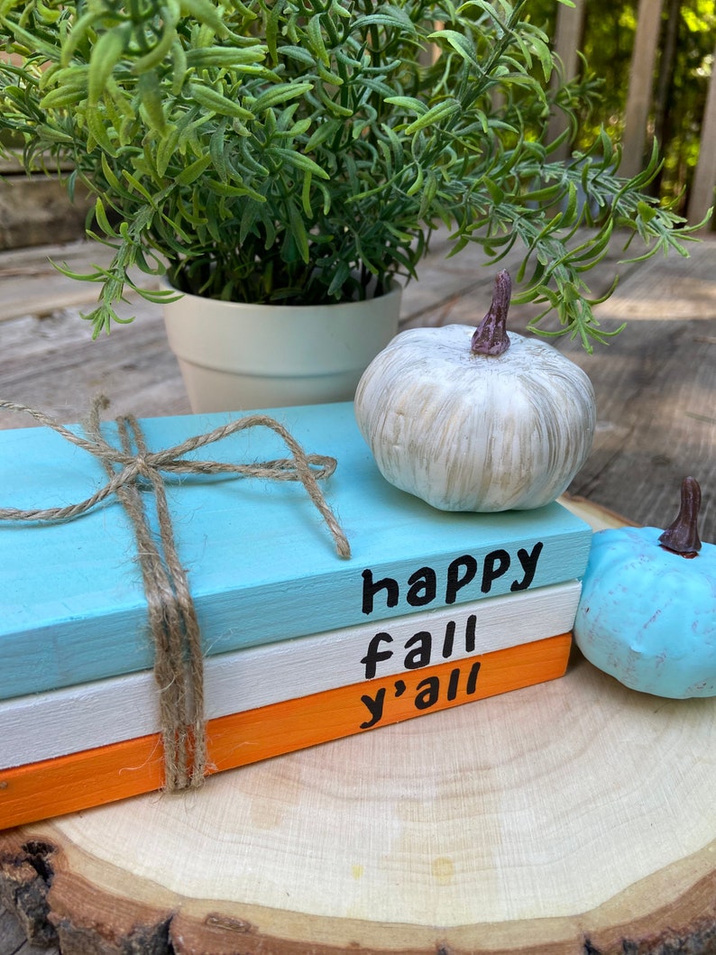 Puede incluir: Tres bloques de madera apilados pintados en azul, blanco y naranja con las palabras "happy fall y'all" escritas en negro. Los bloques est&aacute;n atados con cuerda y est&aacute;n sobre una rodaja de madera con dos peque&ntilde;as calabazas blancas y azules.