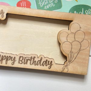 May include: A wooden picture frame with the words "Happy Birthday" engraved along the bottom edge. The frame has a photo aperture and a laser-cut design of balloons on the right. The frame is made of light-coloured wood.