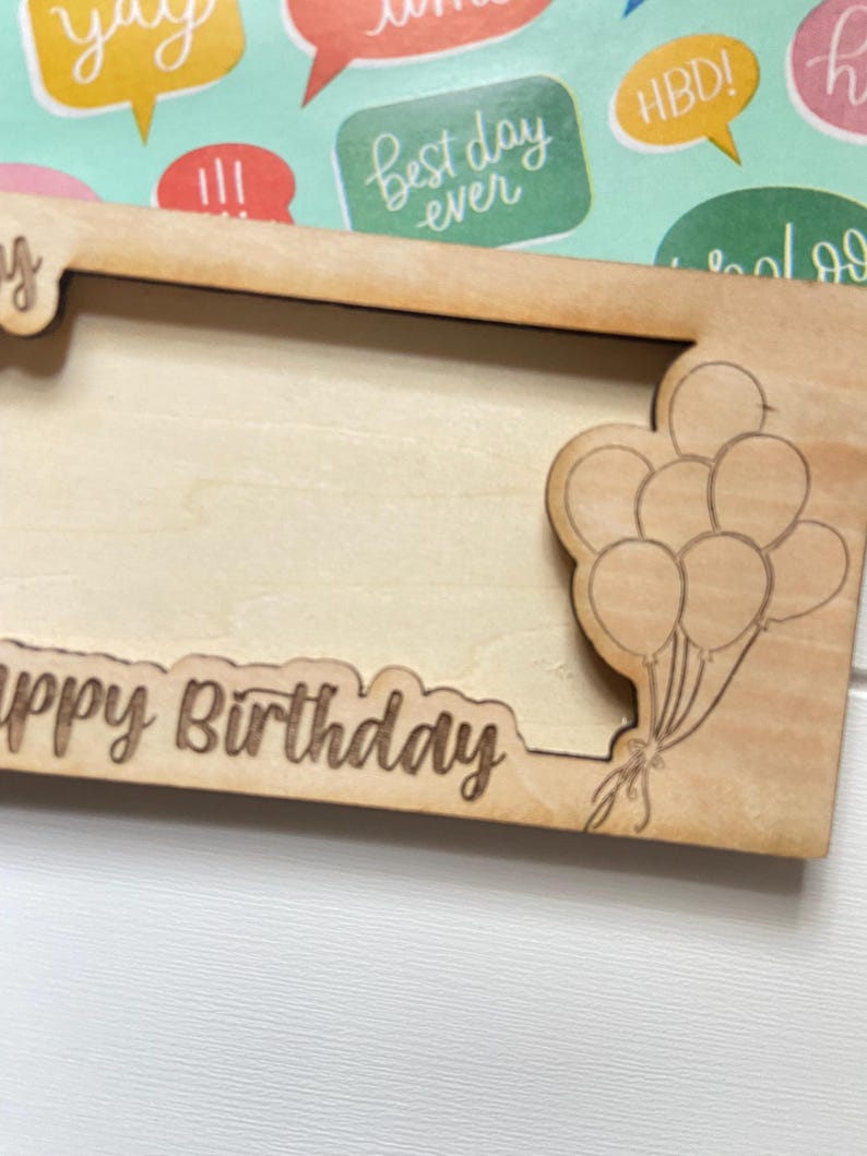May include: A wooden picture frame with the words "Happy Birthday" engraved along the bottom edge. The frame has a photo aperture and a laser-cut design of balloons on the right. The frame is made of light-coloured wood.