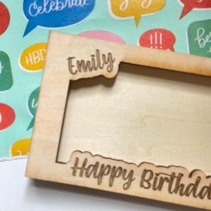 May include: A light-coloured wooden picture frame with the name "Emily" cut out at the top and the words "Happy Birthday" cut out at the bottom. The frame is on a colourful background with speech bubbles.