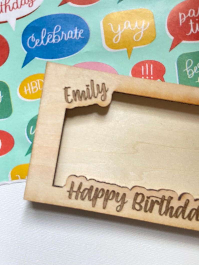May include: A light-coloured wooden picture frame with the name "Emily" cut out at the top and the words "Happy Birthday" cut out at the bottom. The frame is on a colourful background with speech bubbles.