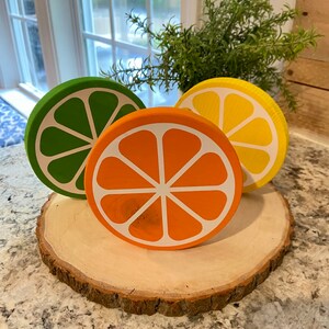 Puede incluir: Tres rodajas de fruta c&iacute;trica de madera sobre una rodaja de madera. Las rodajas est&aacute;n pintadas en tonos de verde, naranja y amarillo. Las rodajas de fruta est&aacute;n dispuestas en c&iacute;rculo sobre la rodaja de madera.