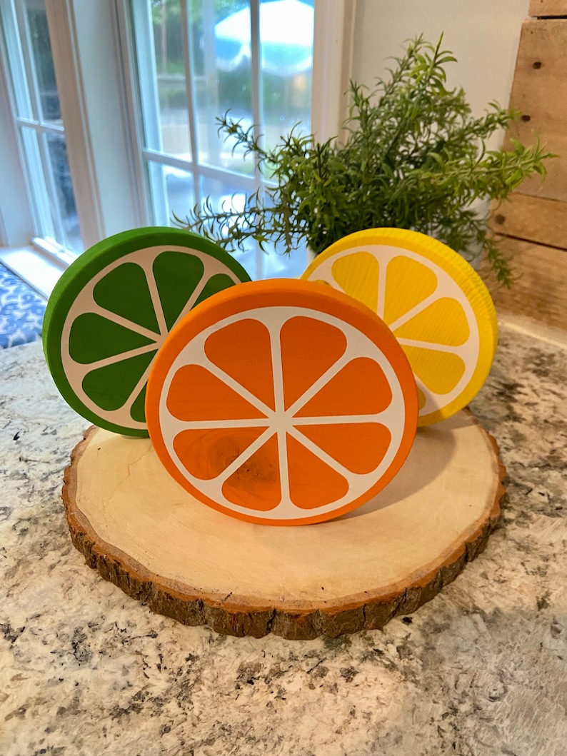 Puede incluir: Tres rodajas de fruta c&iacute;trica de madera sobre una rodaja de madera. Las rodajas est&aacute;n pintadas en tonos de verde, naranja y amarillo. Las rodajas de fruta est&aacute;n dispuestas en c&iacute;rculo sobre la rodaja de madera.