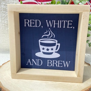 Pu&ograve; includere: Un cartello in legno incorniciato con sfondo blu e testo bianco che recita "Red, White, and Brew". Sopra il testo &egrave; raffigurata una tazza di caff&egrave; bianca con le stelle.