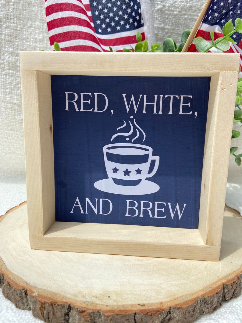 Pu&ograve; includere: Un cartello in legno incorniciato con sfondo blu e testo bianco che recita "Red, White, and Brew". Sopra il testo &egrave; raffigurata una tazza di caff&egrave; bianca con le stelle.