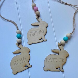K&ouml;nnte beinhalten: Drei h&ouml;lzerne Hasen-Anh&auml;nger mit den Namen "Brinn", "Rory" und "Patrick" eingraviert. Jeder Hase ist an einer Schnur mit Holzperlen und einer rosa oder blauen Perle befestigt.