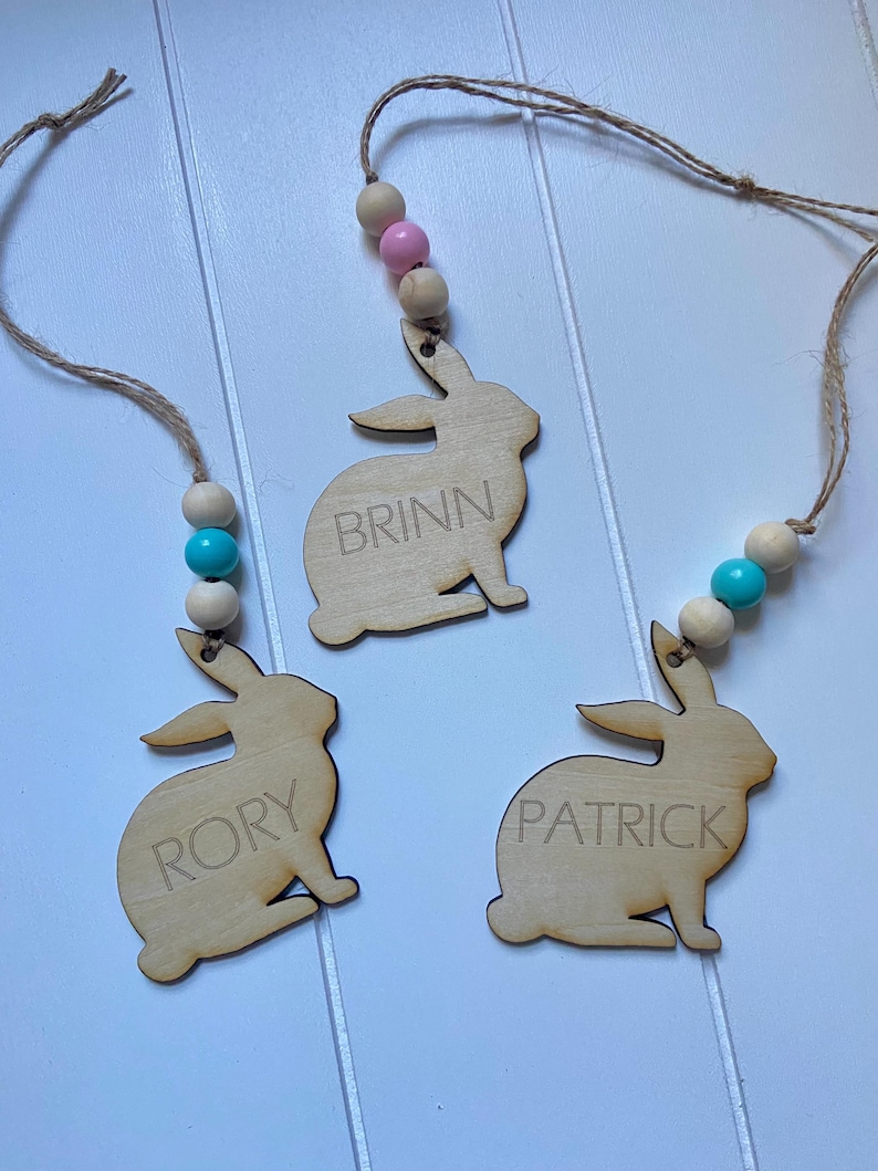 K&ouml;nnte beinhalten: Drei h&ouml;lzerne Hasen-Anh&auml;nger mit den Namen "Brinn", "Rory" und "Patrick" eingraviert. Jeder Hase ist an einer Schnur mit Holzperlen und einer rosa oder blauen Perle befestigt.
