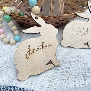 K&ouml;nnte beinhalten: Zwei h&ouml;lzerne Hasenformen mit den Namen "Jonathan" und "Sam" eingraviert. Die Hasen h&auml;ngen an einer Schnur aus pastellfarbenen Holzperlen.