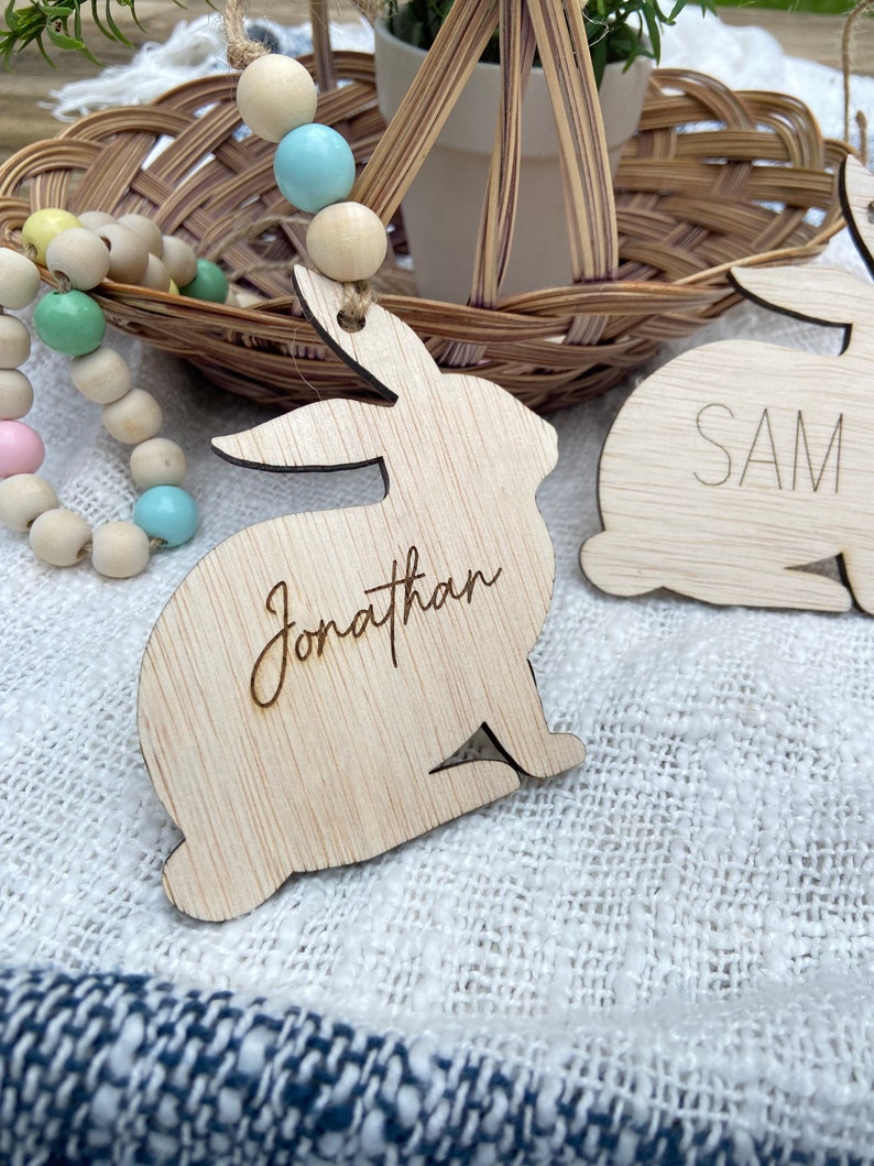 K&ouml;nnte beinhalten: Zwei h&ouml;lzerne Hasenformen mit den Namen "Jonathan" und "Sam" eingraviert. Die Hasen h&auml;ngen an einer Schnur aus pastellfarbenen Holzperlen.