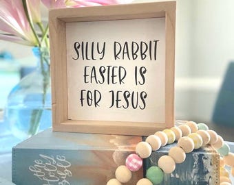 Op de afbeelding: Een houten bord met de tekst "Silly Rabbit Easter is for Jesus" in zwarte letters. Het bord staat op een boek met een blauw en wit bloemenontwerp en een touwtje met houten kralen met roze, groene en gele accenten.