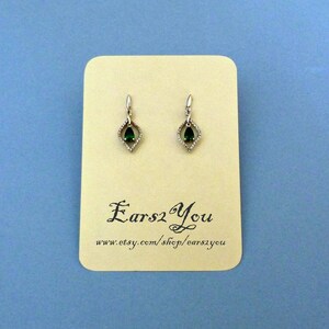 May include: A pair of silver dangle earrings with a teardrop-shaped green gemstone set in a diamond-shaped frame. The earrings are displayed on a card with the text "Ears2You" and a website address.