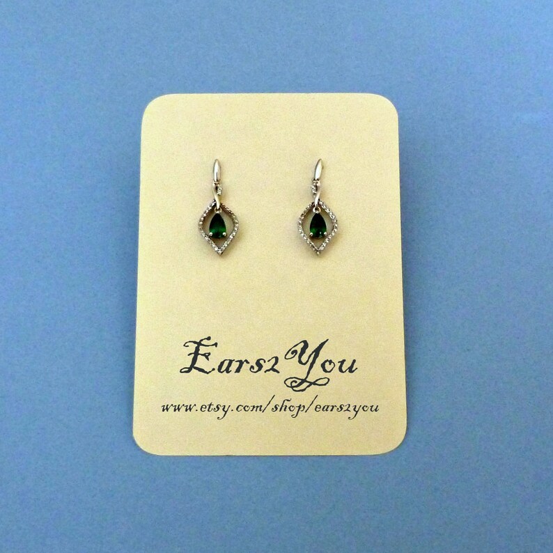 May include: A pair of silver dangle earrings with a teardrop-shaped green gemstone set in a diamond-shaped frame. The earrings are displayed on a card with the text "Ears2You" and a website address.