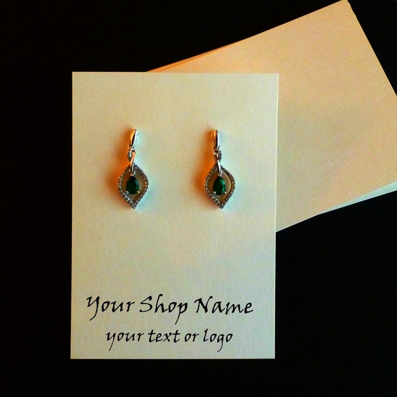 May include: A pair of silver dangle earrings with a green pear-shaped gemstone set in a diamond-shaped frame. The earrings are displayed on a white card with the text "Your Shop Name your text or logo".