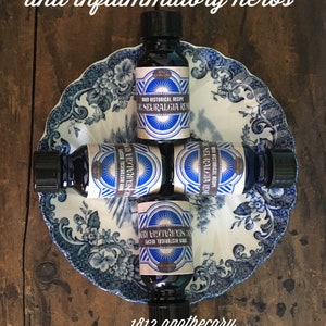 May include: Four small glass bottles with blue and gold labels that read "1889 Historical Recipe Neuralgia Remedy" are arranged in a cross shape on a blue and white porcelain plate. The bottles are on a wooden surface. The text "1812 apothecary Second Chance Farm Sanctuary" is below the plate.