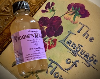Könnte beinhalten: Eine klare Glasflasche mit schwarzem Deckel und lila Etikett mit der Aufschrift "VIRGIN'S MILK" und "Lavendel". Die Flasche befindet sich neben einem Vintage-Buch mit dem Titel "The Language of Flowers" und einer floralen Illustration. Im Hintergrund befindet sich ein dekorativer goldener Rahmen.