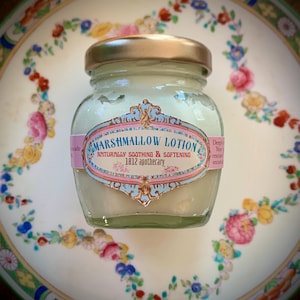 May include: A small glass jar of Marshmallow Lotion with a gold lid, on a decorative plate with floral patterns. The label reads "Marshmallow Lotion, Naturally Soothing & Softening, 1812 apothecary."