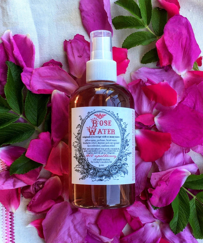 Op de afbeelding: Een doorzichtige glazen fles met een spuitmond, gevuld met rozenwater. De fles heeft een etiket met de tekst "ROSE WATER" en "1812 apothecary" in een vintage lettertype. De fles is omringd door roze rozenblaadjes.