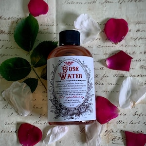 Op de afbeelding: Een doorzichtige glazen fles met een bruine vloeistof erin. De fles is gelabeld met "ROSE WATER" en heeft een vintage ontwerp met een bloemenmotief. De fles staat op een stuk papier met handgeschreven tekst.