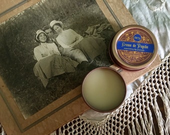 May include: A vintage photo of three people sitting in a field is displayed on a table next to a small tin of Creme de Psyche soothing lip balm. The tin is gold with a blue label and the text "1871 Creme de Psyche soothing lip balm" is printed on the label.