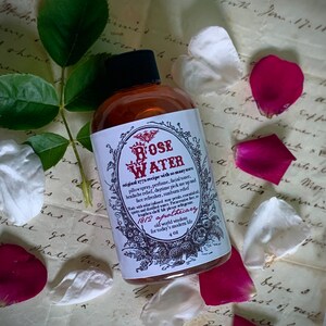 Op de afbeelding: Een glazen flesje met rozenwater met een bruin etiket en gouden letters. Het etiket vermeldt "Rose Water" en "1812 Apothecary". De fles staat op een stuk papier met handschrift.