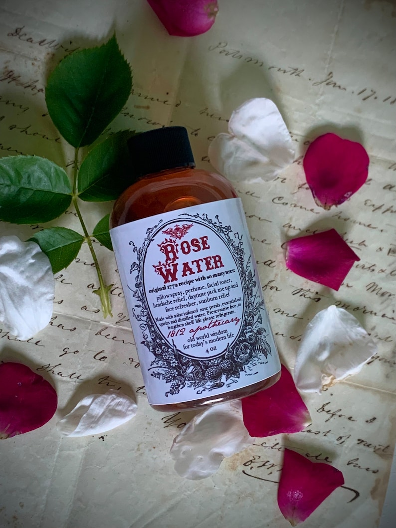 Op de afbeelding: Een glazen flesje met rozenwater met een bruin etiket en gouden letters. Het etiket vermeldt "Rose Water" en "1812 Apothecary". De fles staat op een stuk papier met handschrift.