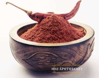 May include: A wooden bowl filled with red chilli powder with a dried red chilli pepper on top. The bowl has a carved design and the text "1812 APOTHECARY" is visible on the bottom.