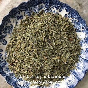 May include: A blue and white floral patterned plate filled with dried green herbs. The plate has a scalloped edge and is set against a light-colored background. Text on the plate reads "1812 apothecary second chance farm sanctuary."
