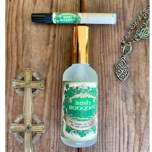 May include: A clear glass bottle with a gold cap and a green label that reads "Irish Bouquet 1812 Apothecary" and "Much admired for its exquisite and long lasting fragrance". The bottle is sitting on a wooden surface with a silver Celtic cross and a silver chain with a Celtic knot pendant.