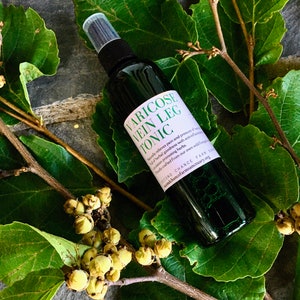 May include: A dark green glass bottle with a white label that reads "Varicose Vein Leg Tonic" and includes information about the product's ingredients and origin. The bottle is sitting on a bed of green leaves.
