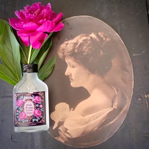 May include: A bottle of Pink Peony Toilette Water with a black cap and a label featuring pink peony flowers. A vibrant pink peony flower and green leaves are next to the bottle. An antique sepia-toned portrait of a woman is in the background.