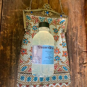 May include: A clear glass bottle with a white label that reads "The Virgin's Milk renders the skin clear and beautiful 1779 An old-fashioned Lotion Ladies use it successfully in every complexion, for which purpose it is better, or indeed for most people, than any other Lotion." The bottle is sitting on a patterned fabric.