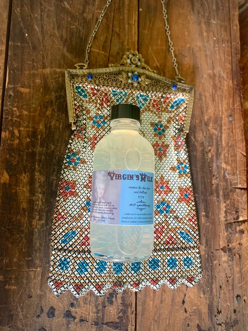May include: A clear glass bottle with a white label that reads "The Virgin's Milk renders the skin clear and beautiful 1779 An old-fashioned Lotion Ladies use it successfully in every complexion, for which purpose it is better, or indeed for most people, than any other Lotion." The bottle is sitting on a patterned fabric.