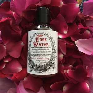 Op de afbeelding: Een doorzichtige glazen fles met rozenwater, een zwarte dop en een wit etiket. Het etiket heeft een vintage ontwerp met de woorden "Rose Water" en "1812 Apothecary". De fles is omringd door roze rozenblaadjes.