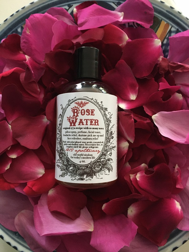 Op de afbeelding: Een doorzichtige glazen fles met rozenwater, een zwarte dop en een wit etiket. Het etiket heeft een vintage ontwerp met de woorden "Rose Water" en "1812 Apothecary". De fles is omringd door roze rozenblaadjes.