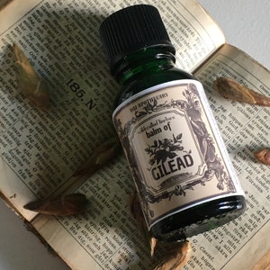 May include: A dark green glass bottle of "Balm of Gilead" tincture, with a cream-colored label featuring black floral illustrations and text. The bottle rests on an open book with dried leaves.