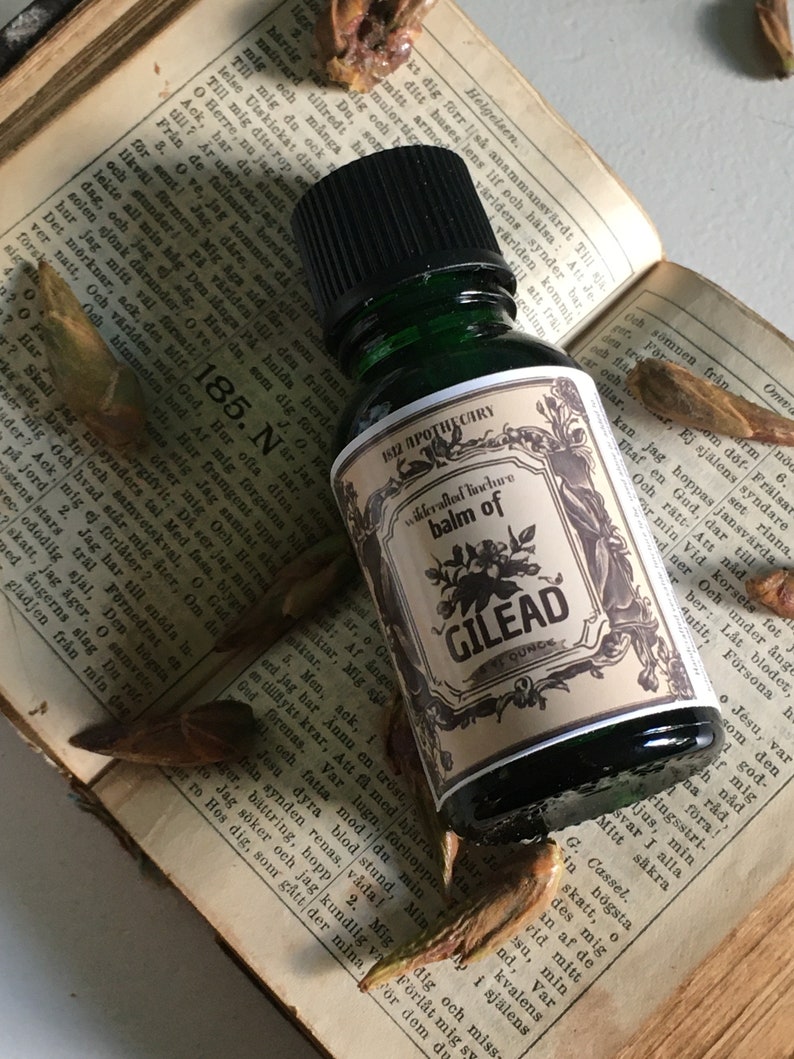 May include: A dark green glass bottle of "Balm of Gilead" tincture, with a cream-colored label featuring black floral illustrations and text. The bottle rests on an open book with dried leaves.