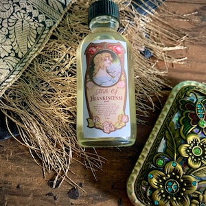 May include: A small glass bottle with a green cap filled with a light yellow liquid. The label reads "Milk of Frankincense" and lists ingredients including frankincense, myrrh, benzoin, sandalwood, bay oil, cedarwood, patchouli, sweet almond oil, and witch hazel. The bottle is sitting on a wooden surface with a decorative gold and green metal object in the background.