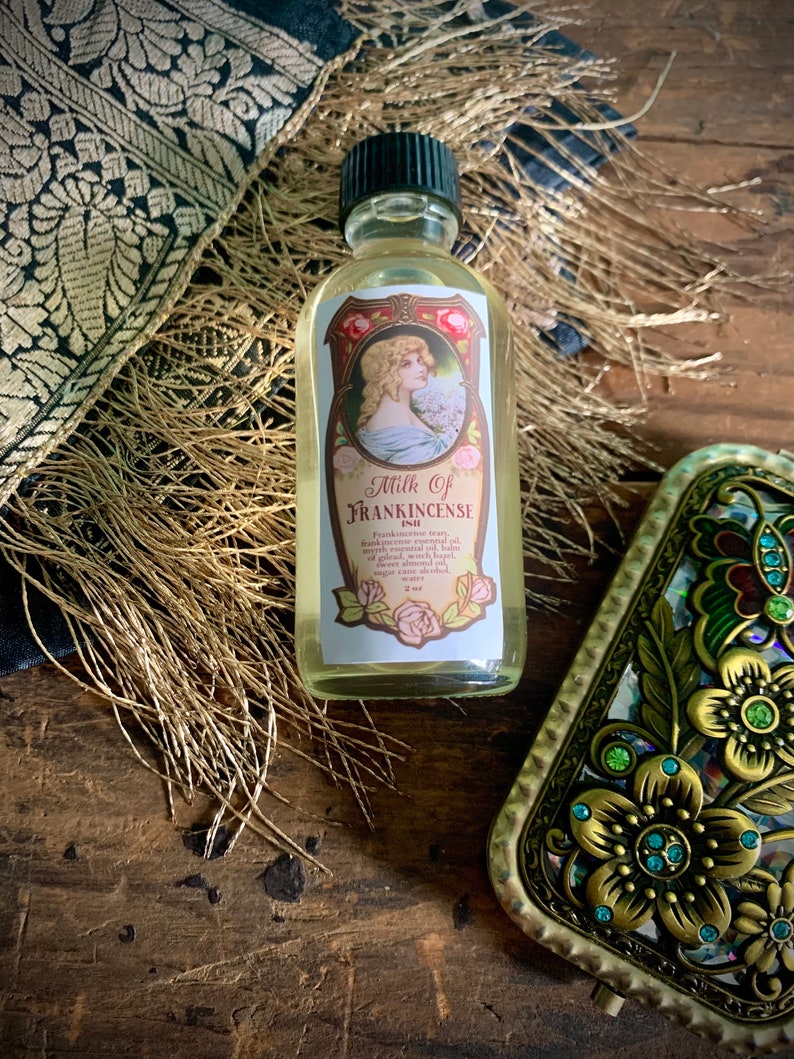 May include: A small glass bottle with a green cap filled with a light yellow liquid. The label reads "Milk of Frankincense" and lists ingredients including frankincense, myrrh, benzoin, sandalwood, bay oil, cedarwood, patchouli, sweet almond oil, and witch hazel. The bottle is sitting on a wooden surface with a decorative gold and green metal object in the background.
