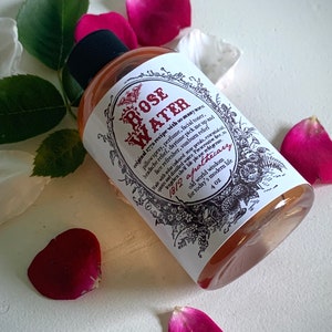 Op de afbeelding: Een doorzichtige glazen fles met een wit etiket waarop "ROSE WATER" staat en veel toepassingen voor het product worden vermeld. Het etiket bevat ook de tekst "1812 apothecary" en "4 oz". De fles is omringd door roze rozenblaadjes.