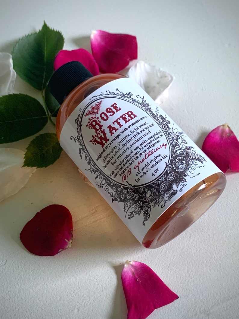 Op de afbeelding: Een doorzichtige glazen fles met een wit etiket waarop "ROSE WATER" staat en veel toepassingen voor het product worden vermeld. Het etiket bevat ook de tekst "1812 apothecary" en "4 oz". De fles is omringd door roze rozenblaadjes.