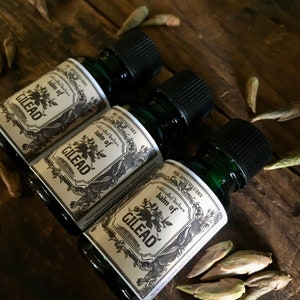 May include: Three dark green glass bottles with black caps, labeled "Balm of Gilead" with floral and decorative elements. The bottles are arranged on a dark wooden surface, surrounded by small, dried seed pods.