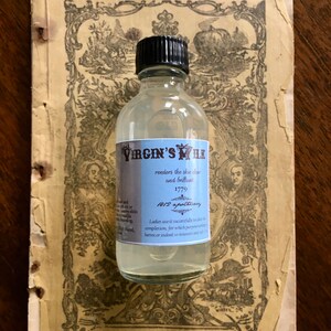 May include: A clear glass bottle with a black cap labeled "Virgin's Milk" and the text "renders the skin clear and brilliant 1779 1812 apothecary Ladies use it successfully to render the complexion, for which purpose nothing is better, or indeed so innocent and safe." The bottle is sitting on a vintage book with a decorative design.